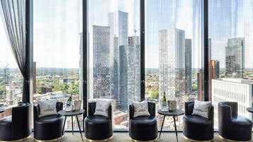 A modern hotel room with large windows, black leather chairs, and a view of the cityscape.