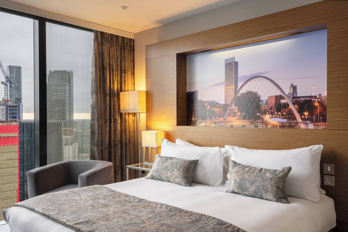 Hotelkamer met groot bed, een stoel, nachtkastje, lamp en stadsgezicht door grote ramen.