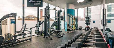 A gym with various exercise equipment, including dumbbells and weight machines, and large windows offering views of the city.