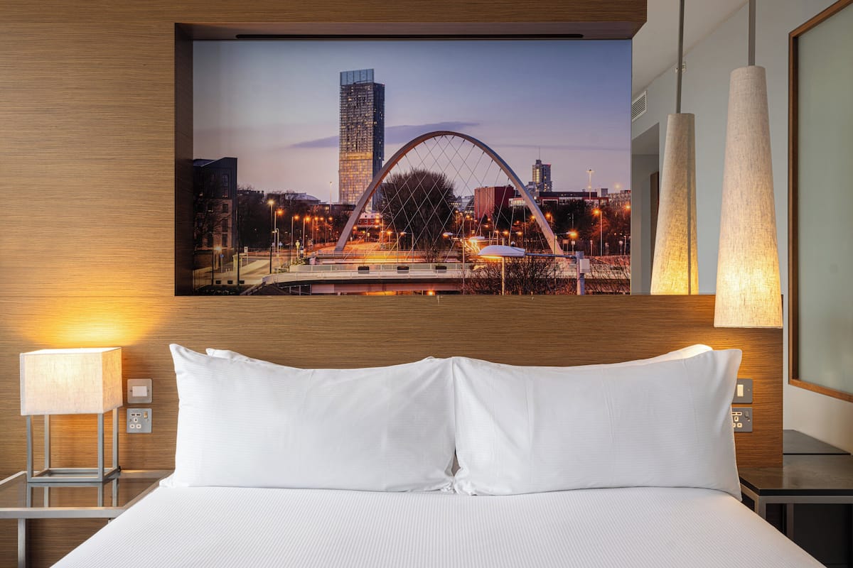 Een hotelkamer met een groot bed, een aan de muur bevestigde televisie waarop een stadsgezicht te zien is, en een nachtkastje met een lamp.