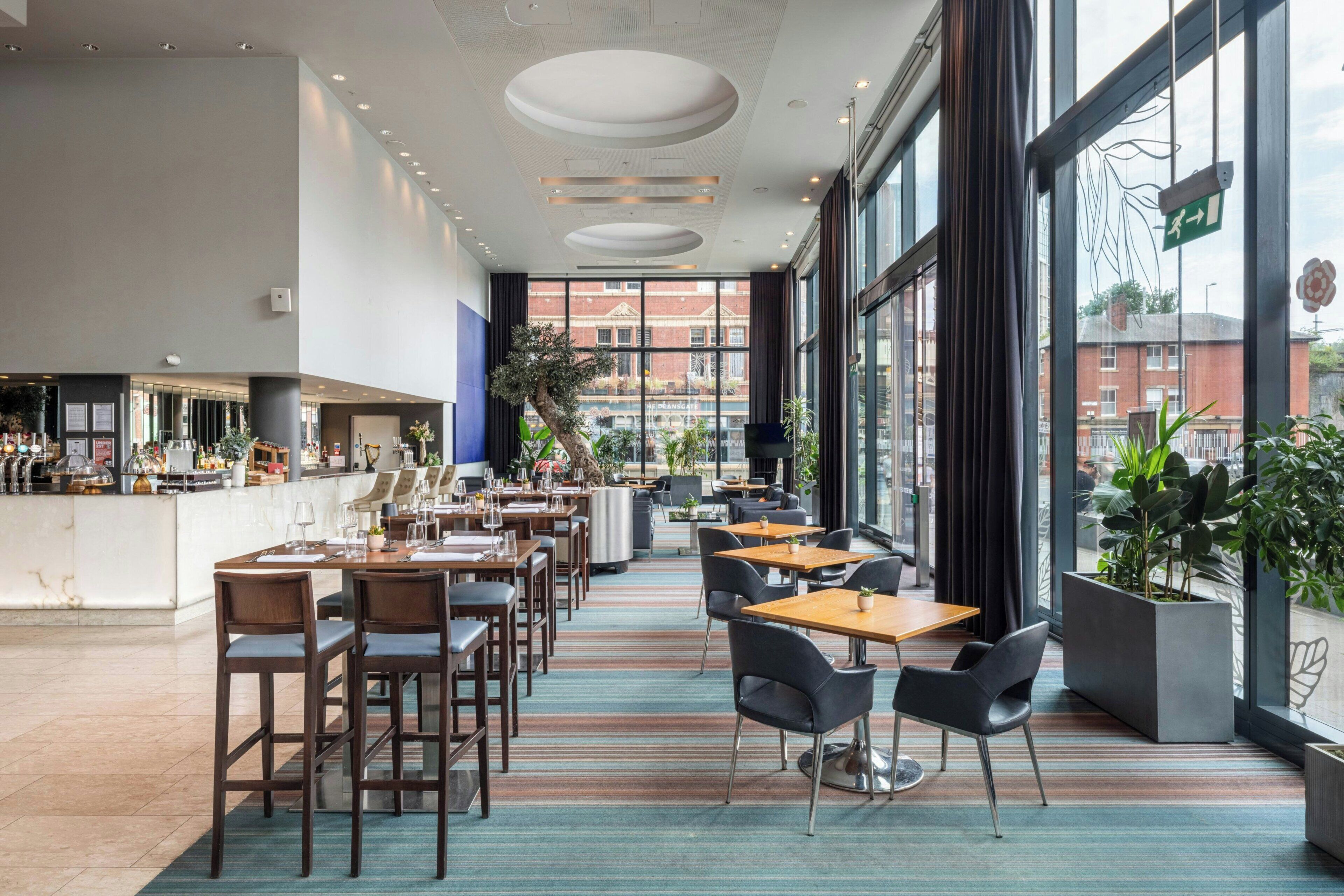 Un restaurante moderno con zona de bar, diversas opciones de asientos y grandes ventanales con vista a la ciudad.