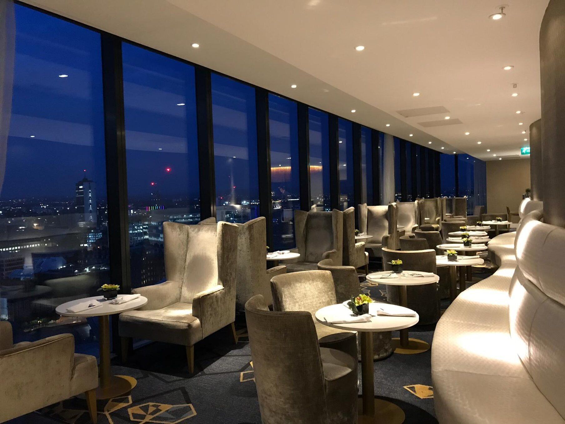 Un moderno salón de hotel con vista a la ciudad, que ofrece asientos cómodos y mesitas.