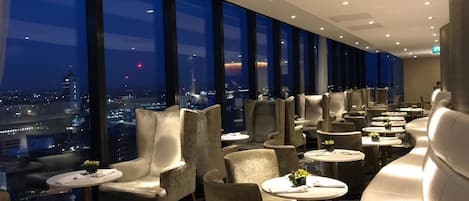 Un moderno salón de hotel con vista a la ciudad, que ofrece asientos cómodos y mesitas.