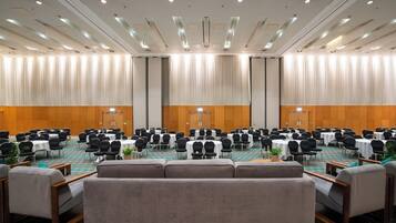 Un salón de conferencias con filas de mesas y sillas, un escenario y una zona de estar con sofás.