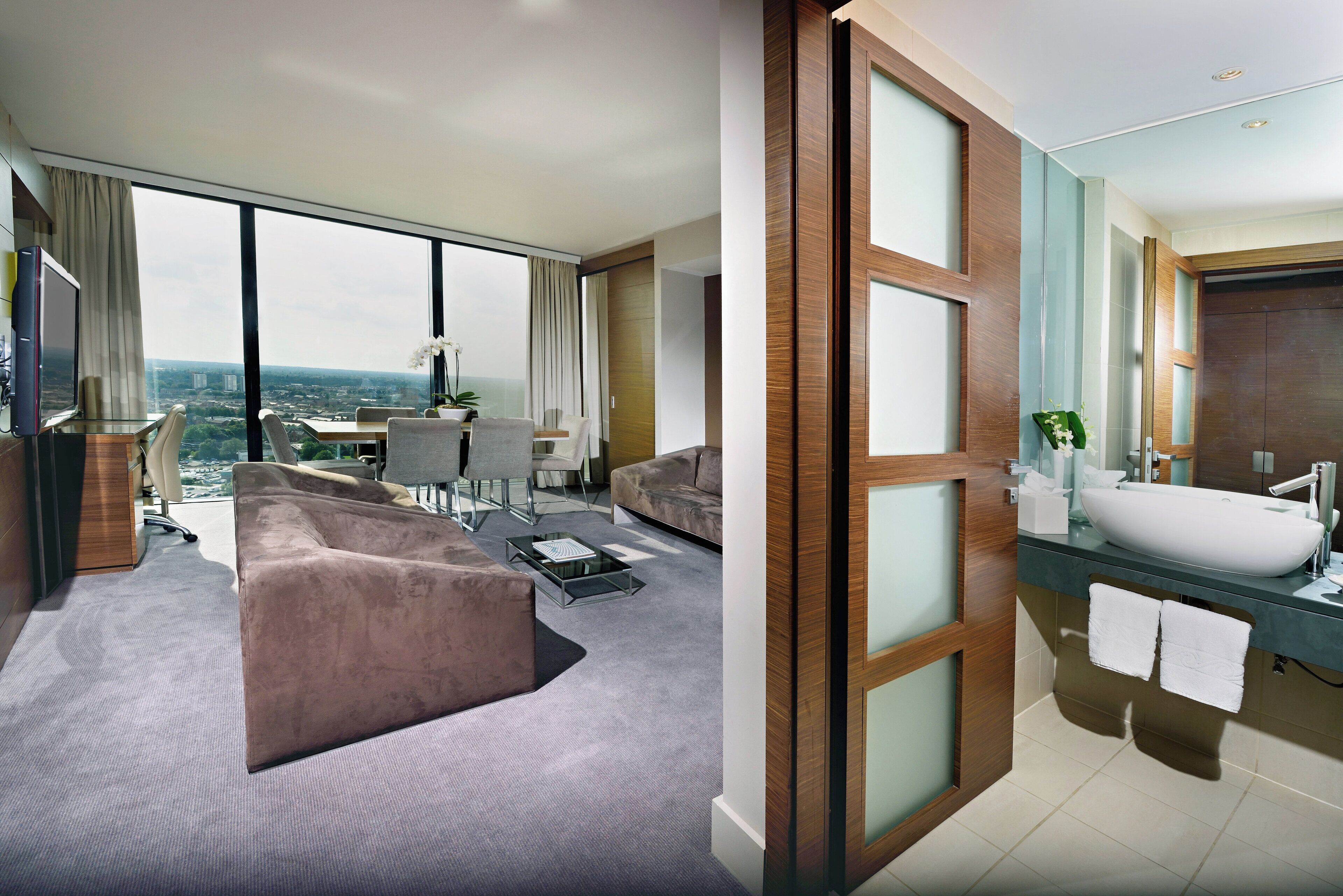 A modern hotel room with a large window, a sofa, a dining area, and a bathroom with a large sink and mirror.