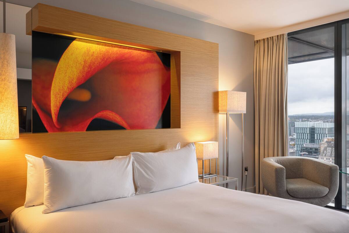 Hotelkamer met een groot bed, uitzicht over de stad, een kunstwerk aan de muur en een nachtkastje met een lamp.