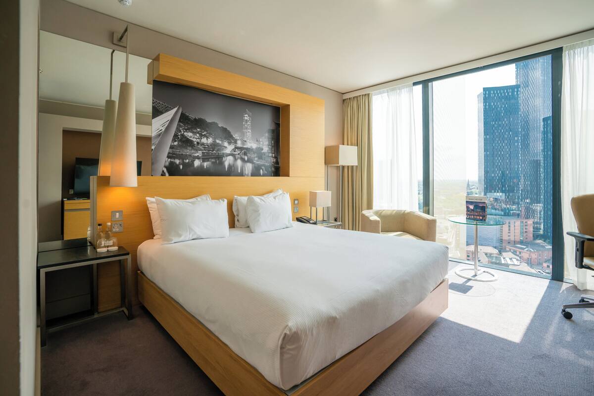Une chambre d’hôtel avec un grand lit, un bureau, une chaise, une grande fenêtre donnant sur la ville et un tableau au mur.