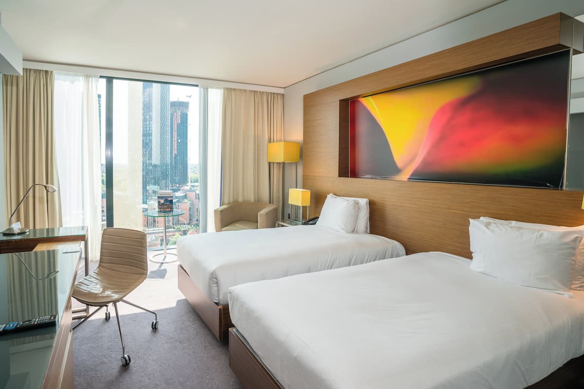Une chambre d’hôtel avec deux lits, un bureau avec une chaise, une lampe et une grande fenêtre donnant sur la ville.