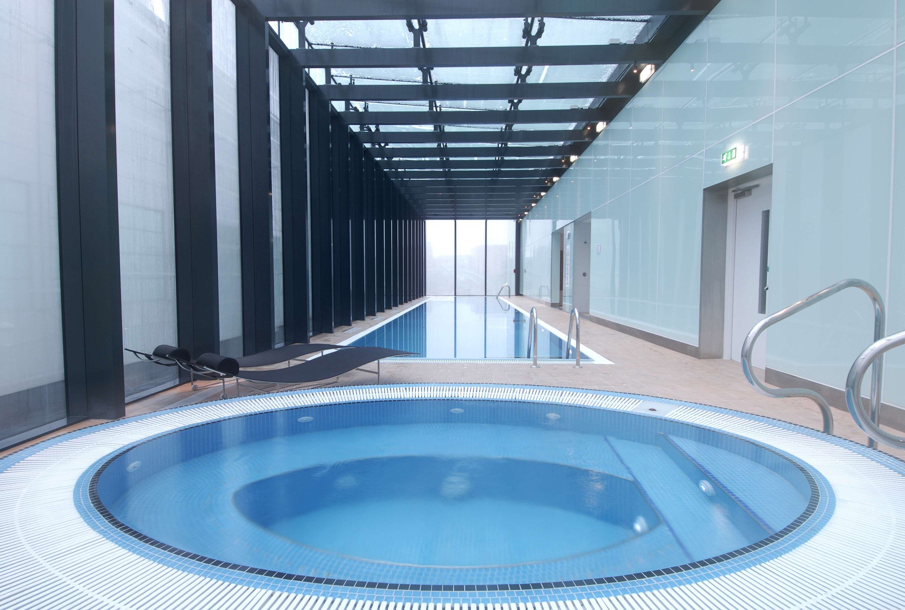 Ein Schwimmbecken mit Fliesenrand, eine Lounge mit Sesseln und ein gläserner Gang.