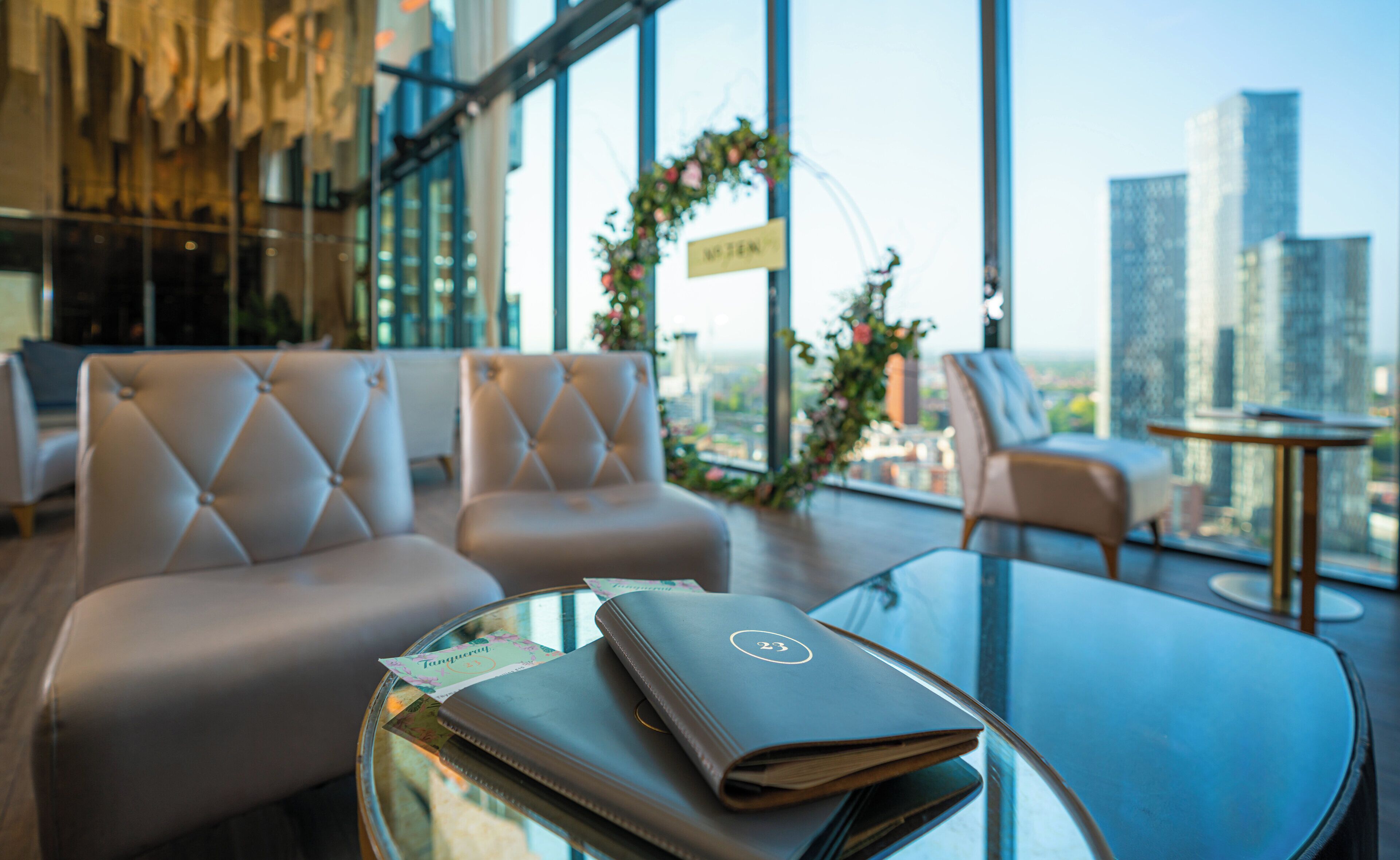 Un hall d’hôtel moderne, doté d’un espace salon, d’une table en verre sur laquelle repose un livre à la couverture en cuir, et offrant une vue sur le paysage urbain grâce à de grandes fenêtres.