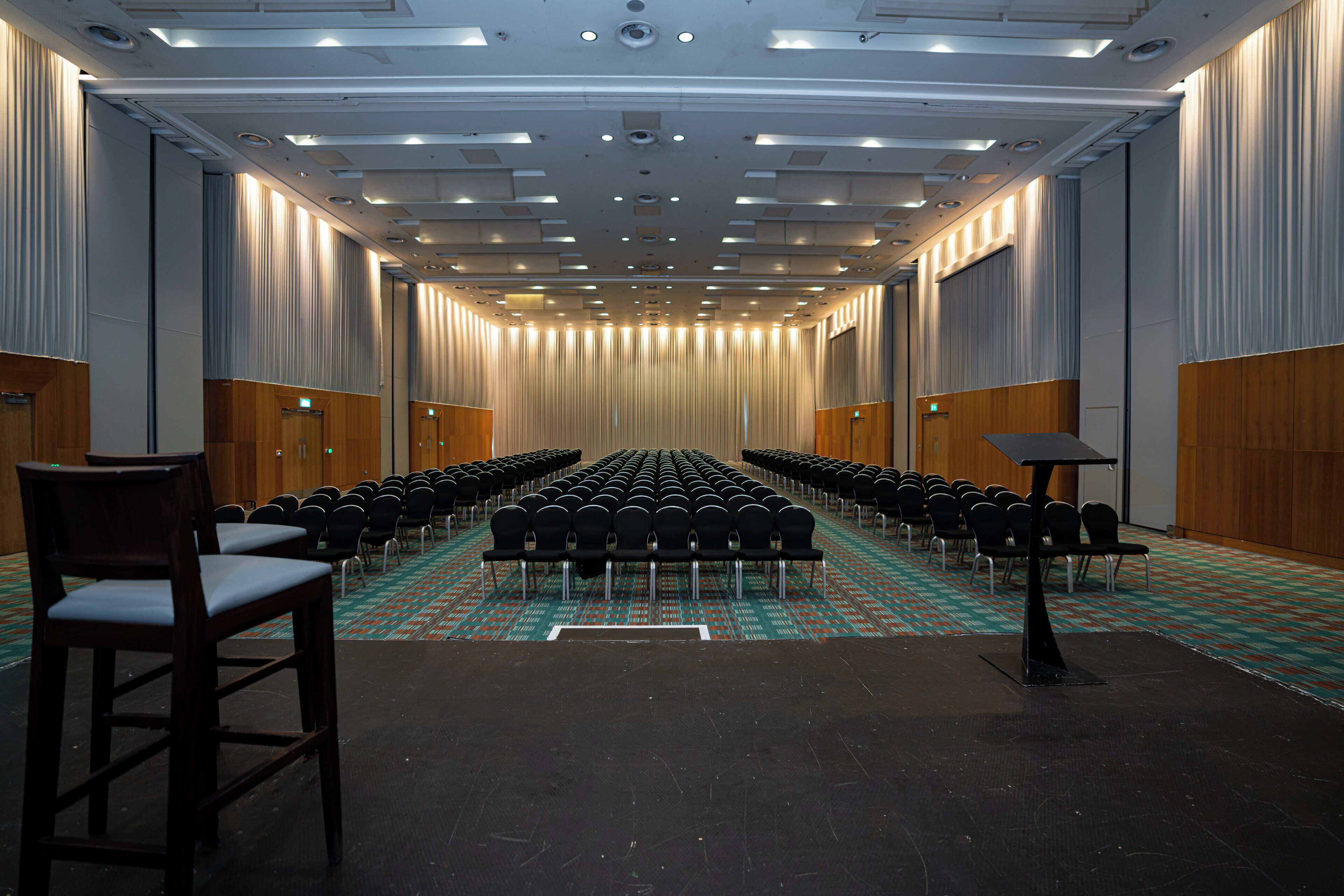 Een vergaderzaal met rijen stoelen, een podium en een vloerbedekking.