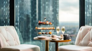 A three-tiered dessert stand with pastries and a glass of champagne on a table in a room with two upholstered chairs and a large window with a cityscape view. - The Manchester Deansgate Hotel by IHG (Manchester)