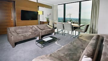 A modern hotel room with a large window, a flat-screen TV, a sofa, a dining table with chairs, and a small coffee table.