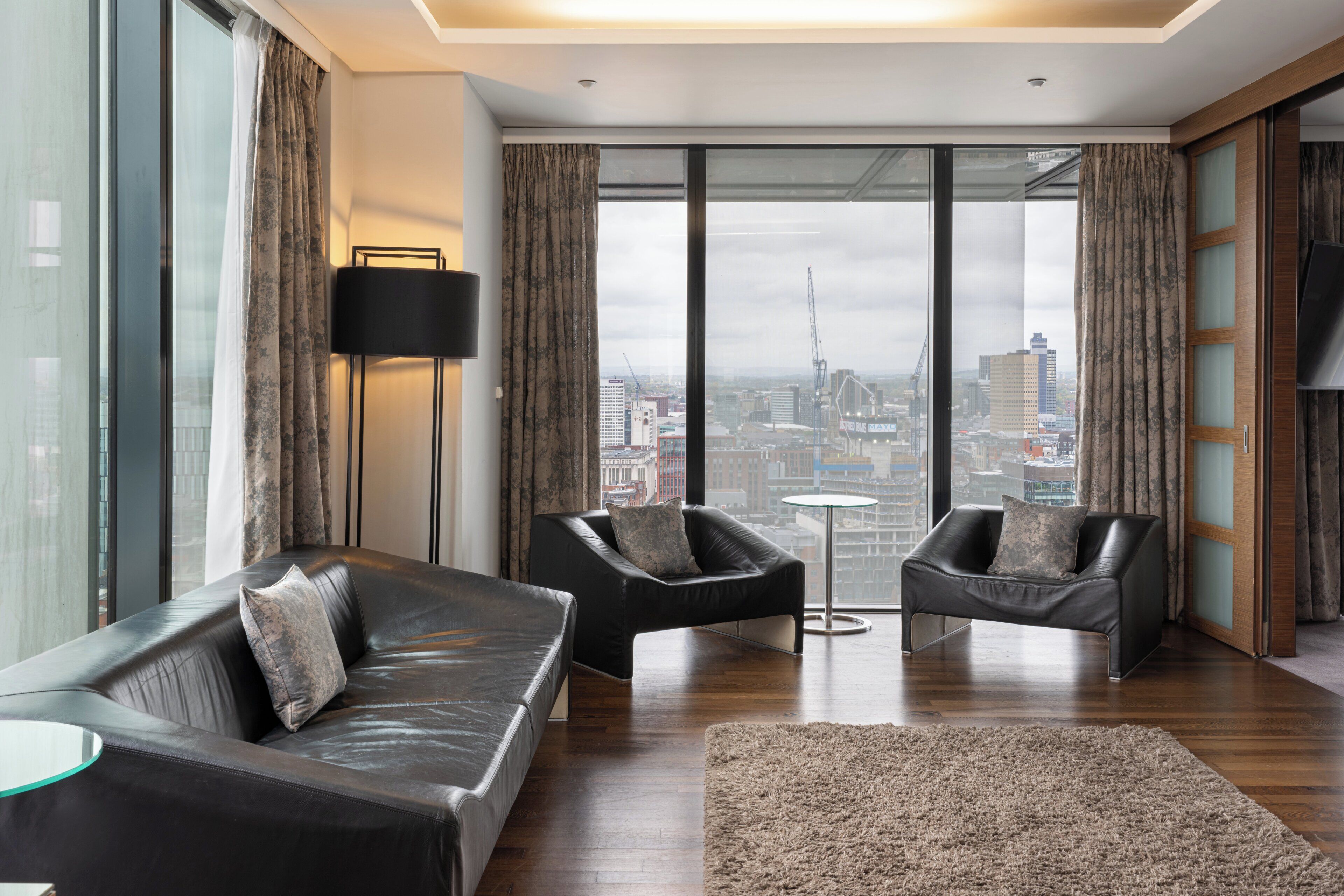A modern living room with a sofa, two armchairs, a small round table, and a large window offering a city view.