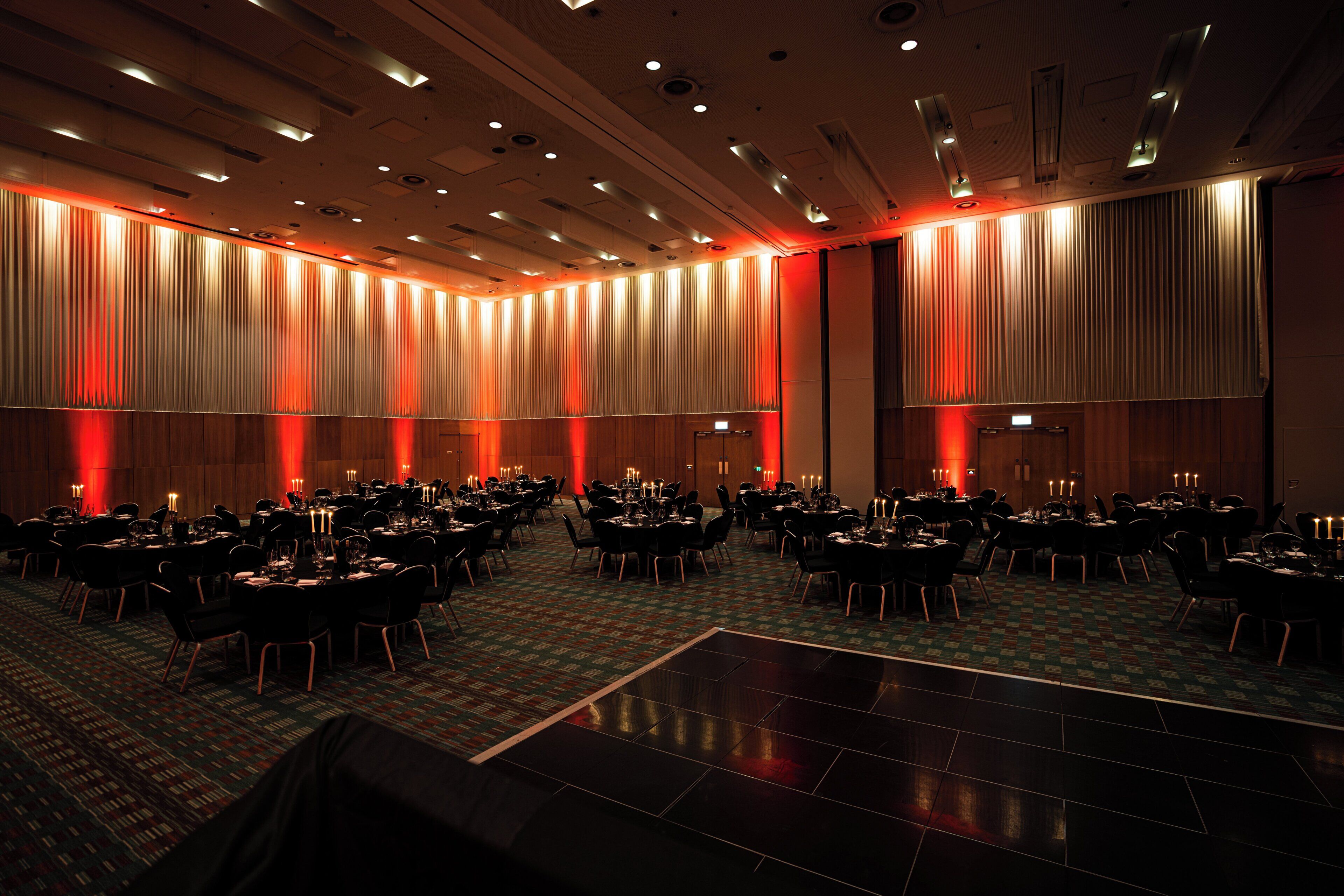 A large banquet hall with multiple round tables and chairs, a patterned carpet, and a high ceiling with recessed lighting.