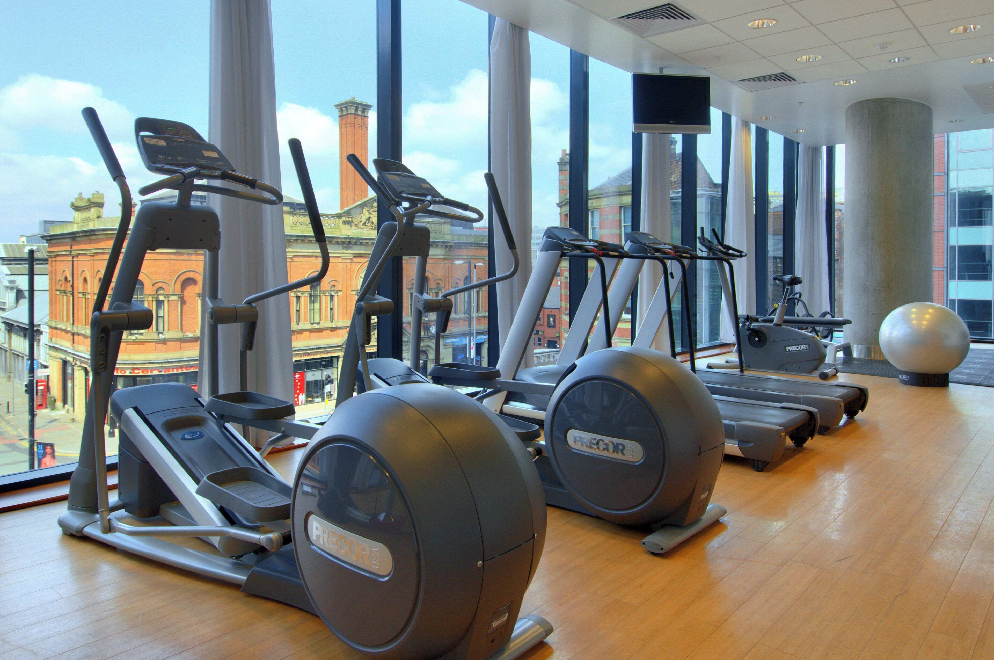 A gym with treadmills, elliptical machines, and a stationary bike, with large windows offering a view of buildings outside.