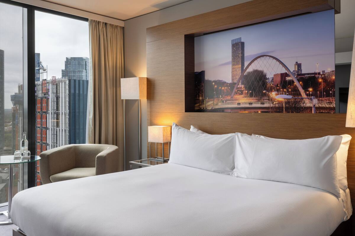 Une chambre d’hôtel avec un grand lit, une chaise, une table en verre, une lampe et une vue sur la ville par la fenêtre.