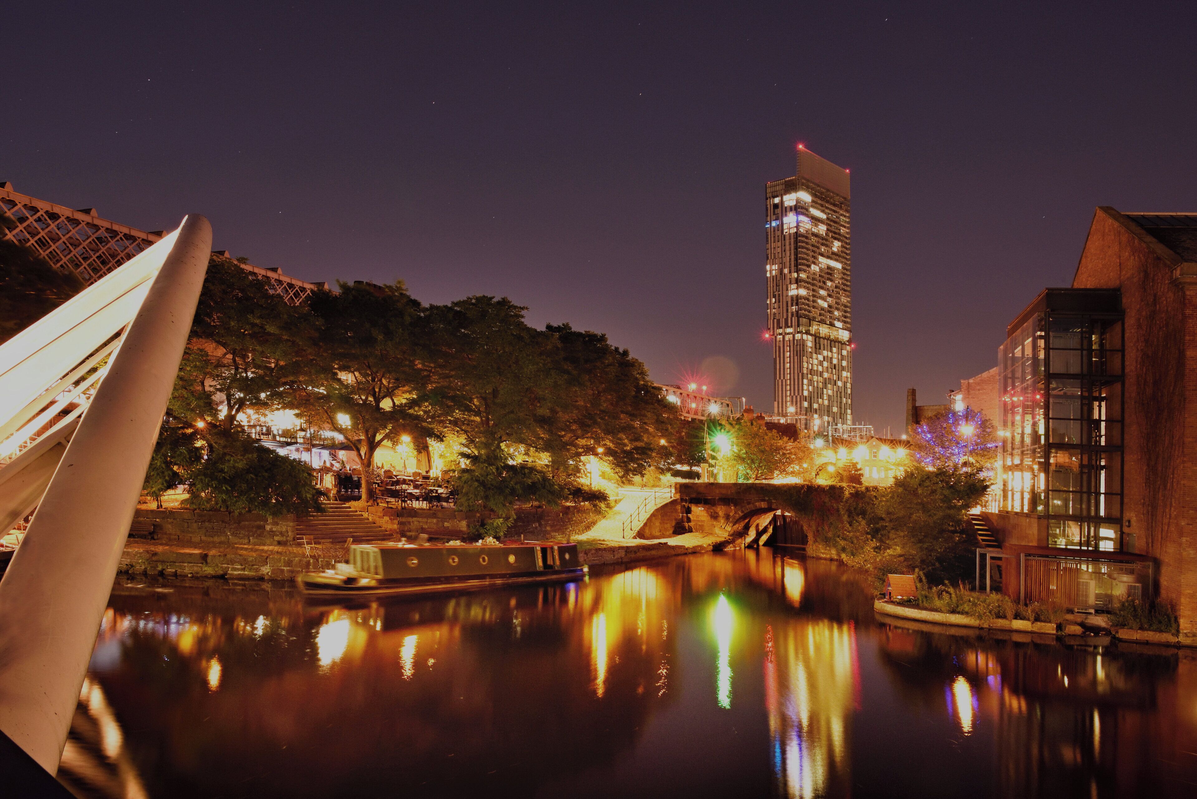 A nighttime cityscape with a river reflecting lights, a modern building, and a bridge.
