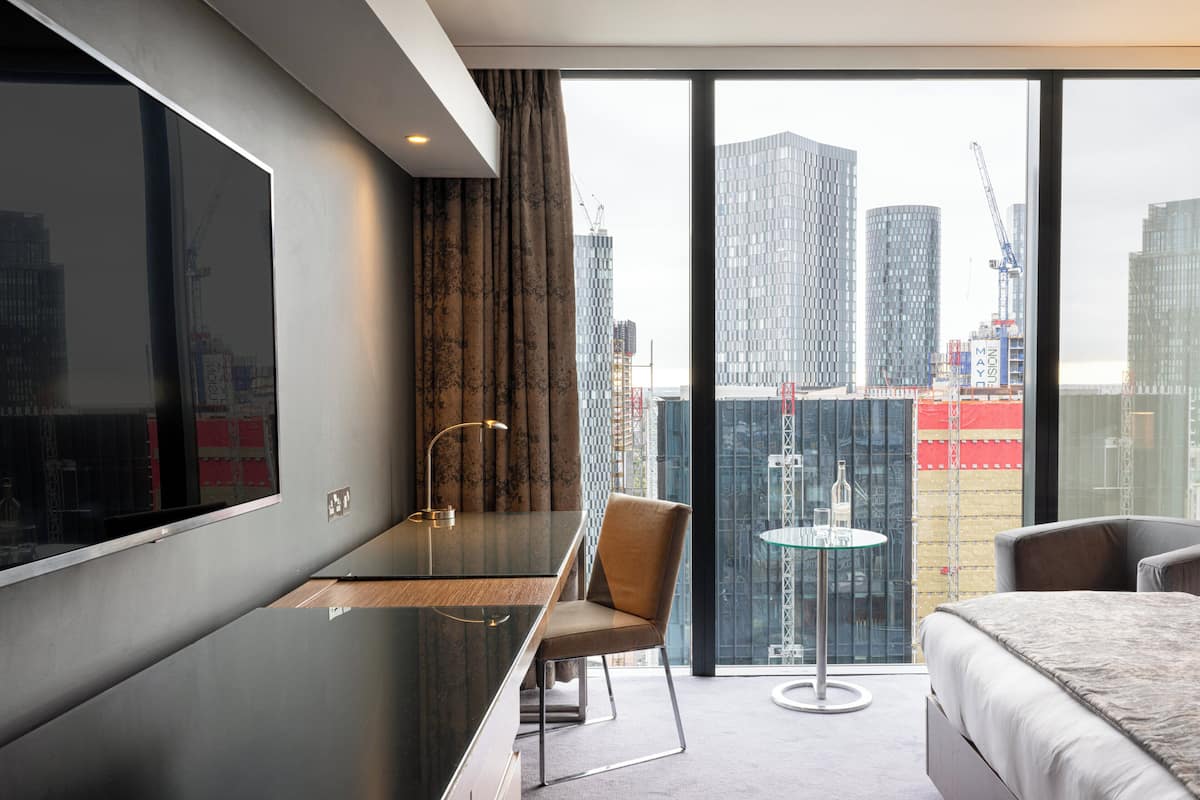Une chambre d’hôtel moderne dotée d’un grand lit, d’un bureau, d’une chaise et offrant une vue sur le paysage urbain grâce à de grandes fenêtres.