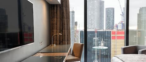 Ein modernes Hotelzimmer mit einem großen Bett, einem Schreibtisch, einem Sessel und Blick auf die Stadtlandschaft durch große Fenster.