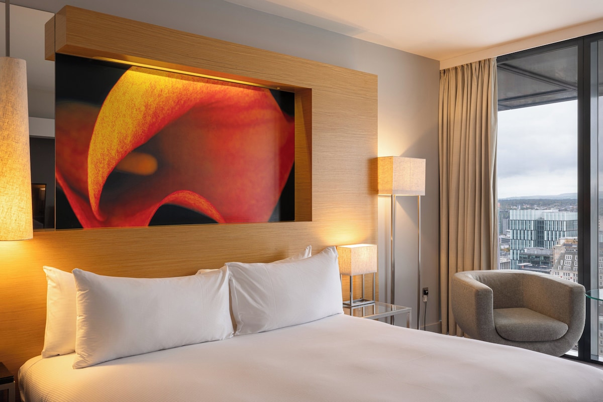 Hotelkamer met een groot bed, uitzicht over de stad, een kunstwerk aan de muur en een nachtkastje met een lamp.
