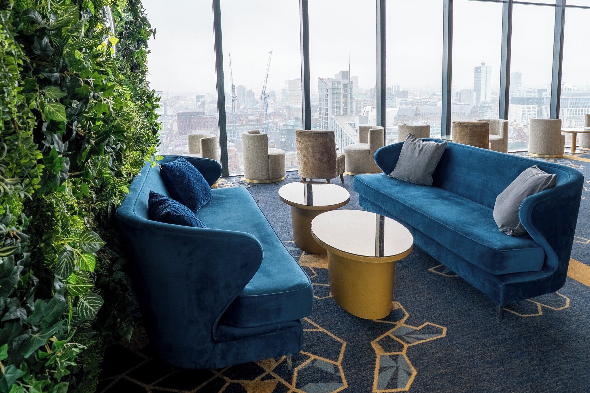 A modern lounge area with a city view, featuring blue sofas, round gold tables, and a green vertical garden.