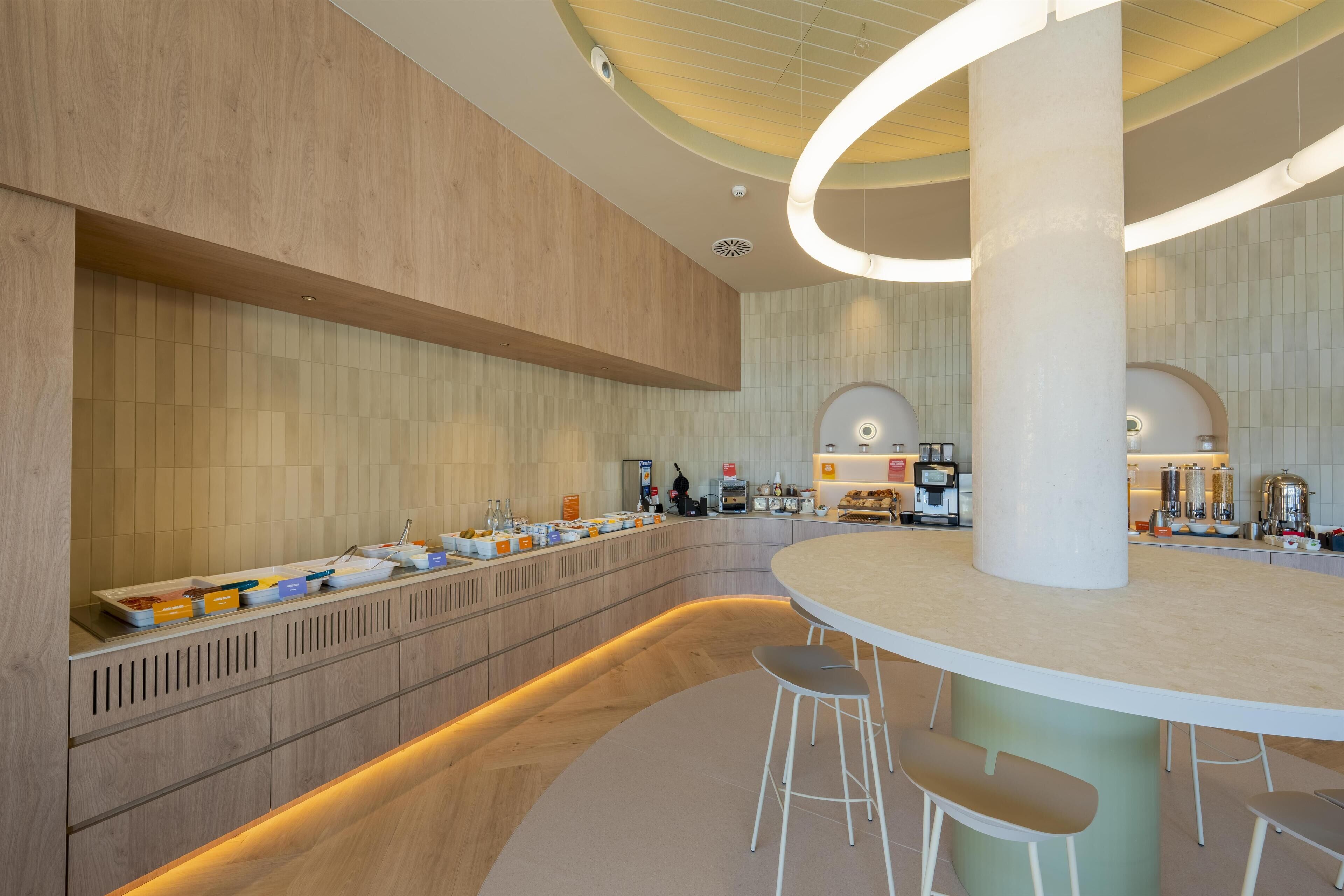A modern hotel lobby with a curved reception desk, a long buffet counter, and a seating area with stools.