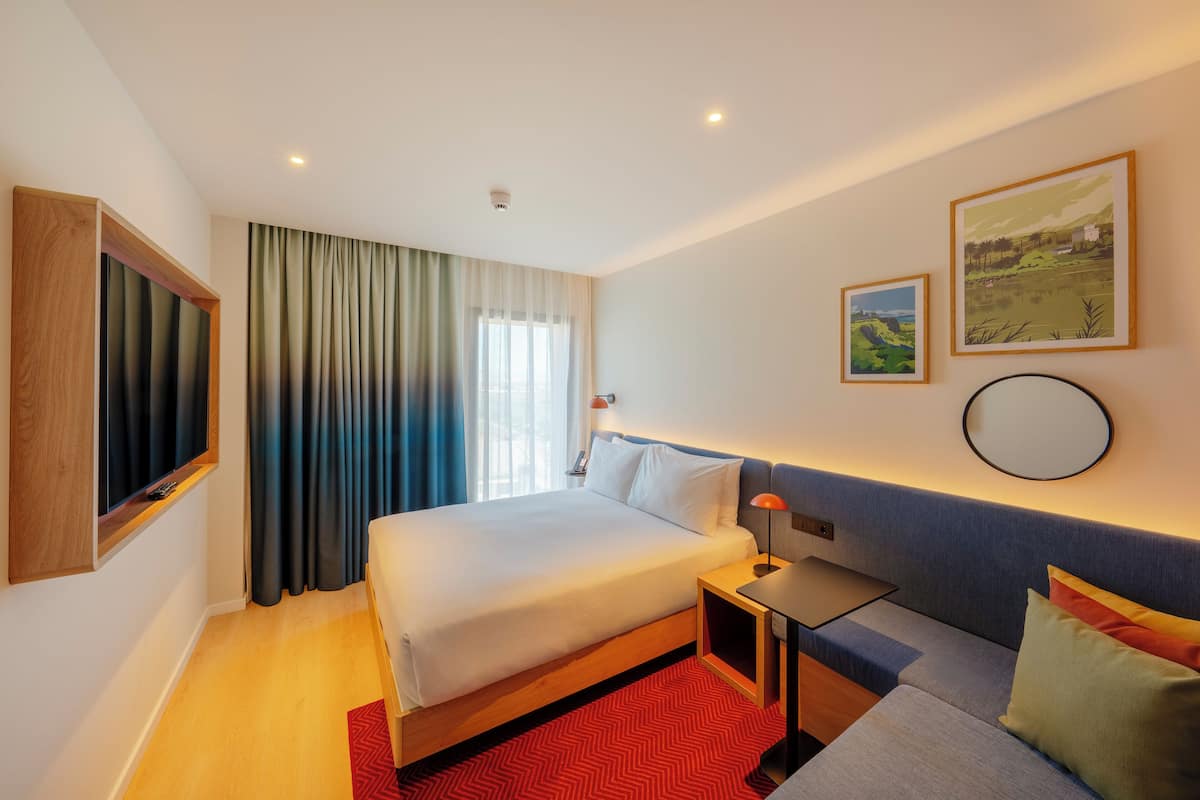 Quarto de hotel moderno com uma cama grande, uma escrivaninha e um sofá. Há duas pinturas na parede e uma janela com cortinas.