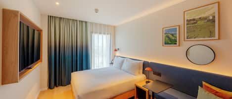 A modern hotel room with a large bed, a desk, and a sofa. There are two paintings on the wall and a window with curtains.