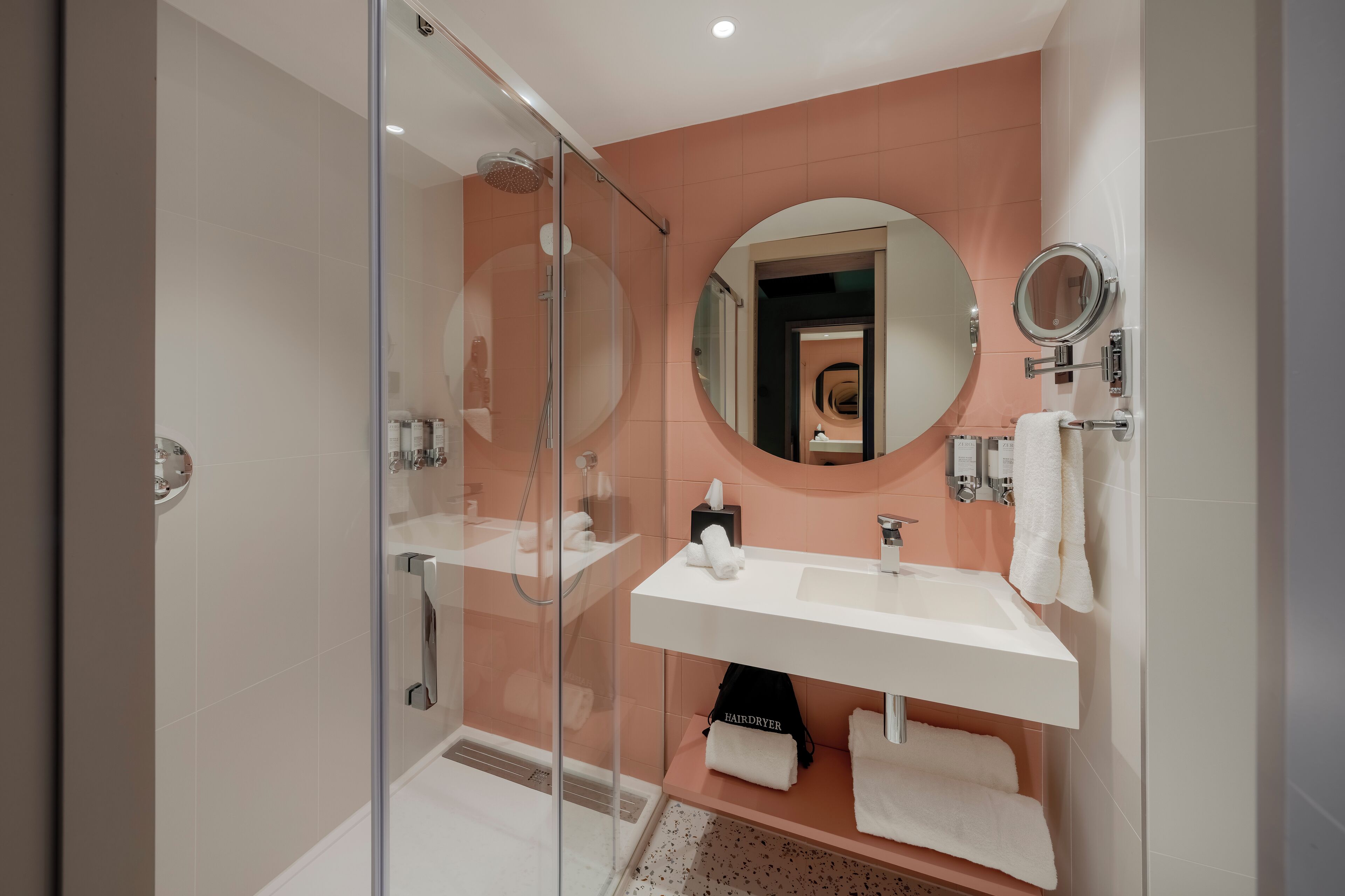 Un baño moderno con ducha de vidrio, lavamanos blanco, espejo redondo y toallero.