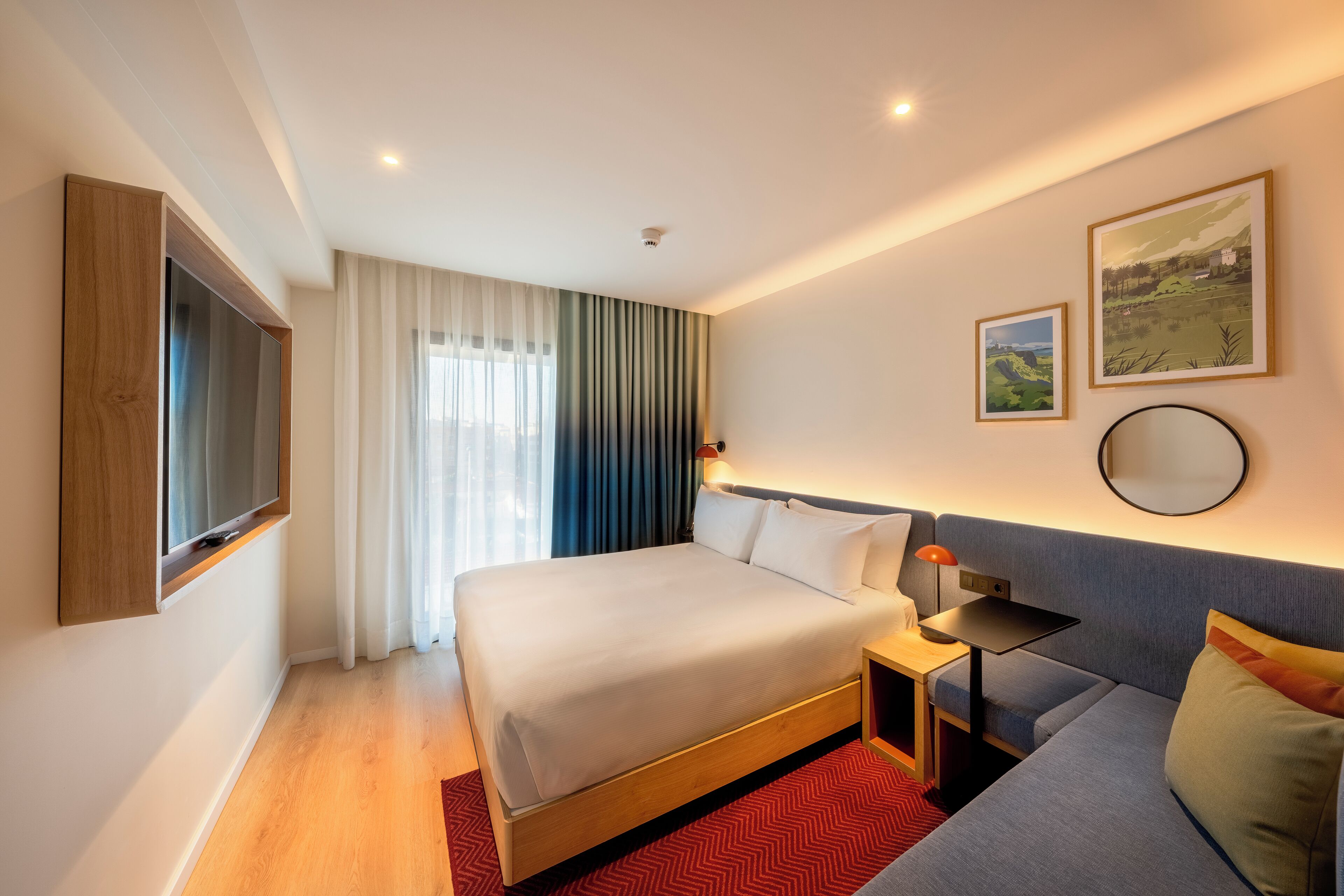 Una habitación de hotel moderna con una cama grande, un sofá, un televisor montado en la pared y dos cuadros enmarcados.