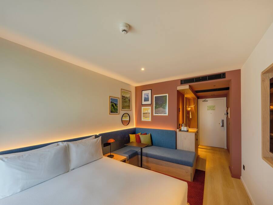 Quarto de hotel com uma cama, sofá, pequena mesa e escrivaninha. Há quadros nas paredes e uma porta que dá para outro espaço.
