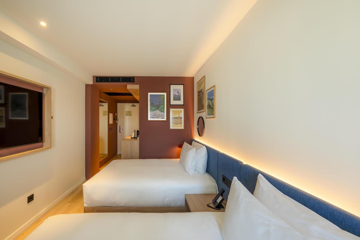 Quarto de hotel com duas camas, uma escrivaninha, um telefone e quadros na parede.