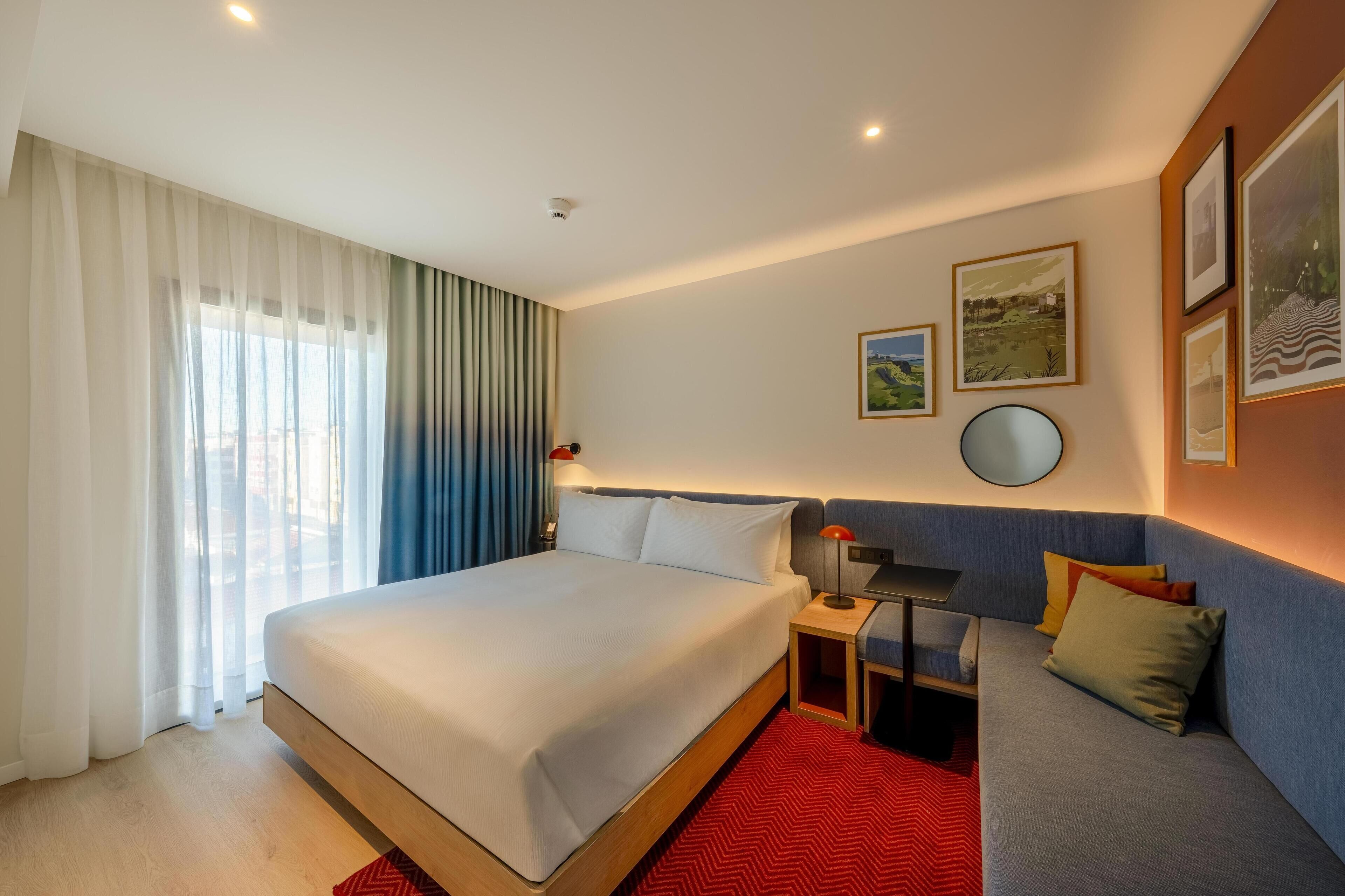 Une chambre d’hôtel avec un grand lit, un canapé, une petite table et un grand miroir rond fixé au mur.