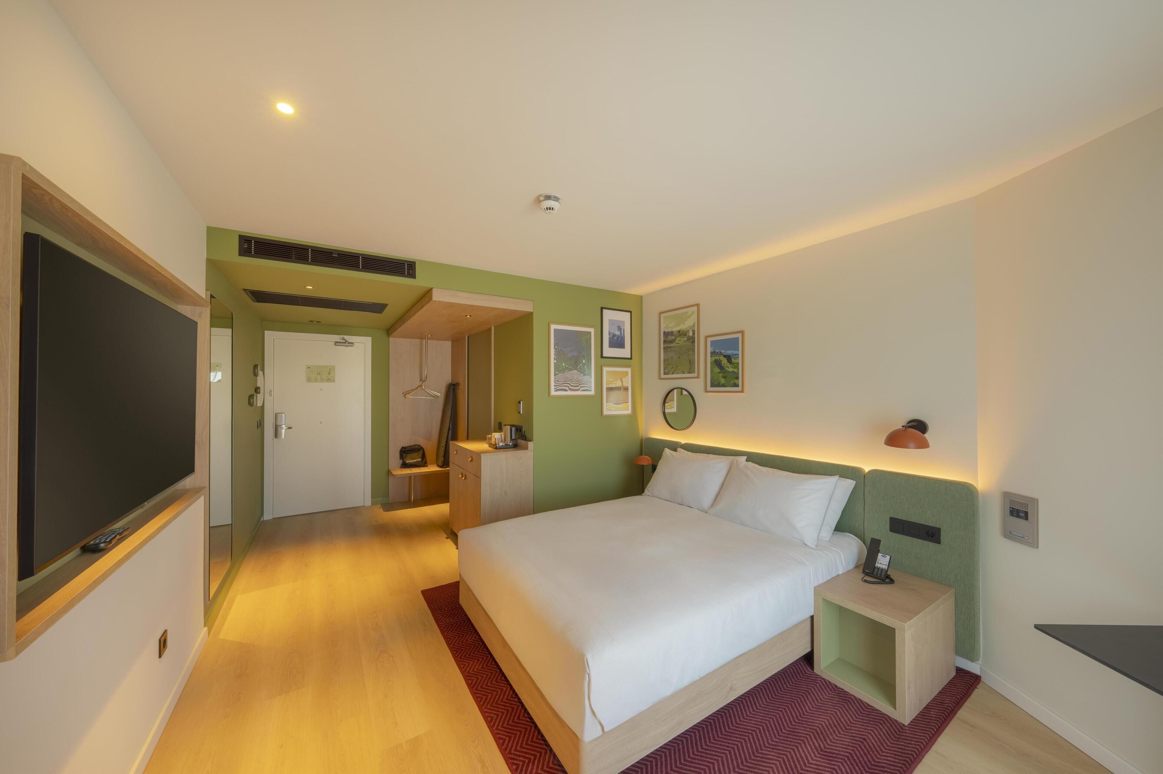 A hotel room with a bed, bedside table, wall-mounted lamp, and a door leading to another room.