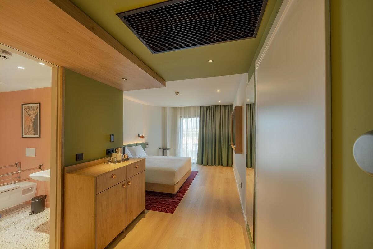 Quarto de hotel moderno com cabeceira de madeira, cama grande, escrivaninha embutida e vista para a cidade através de grandes janelas.