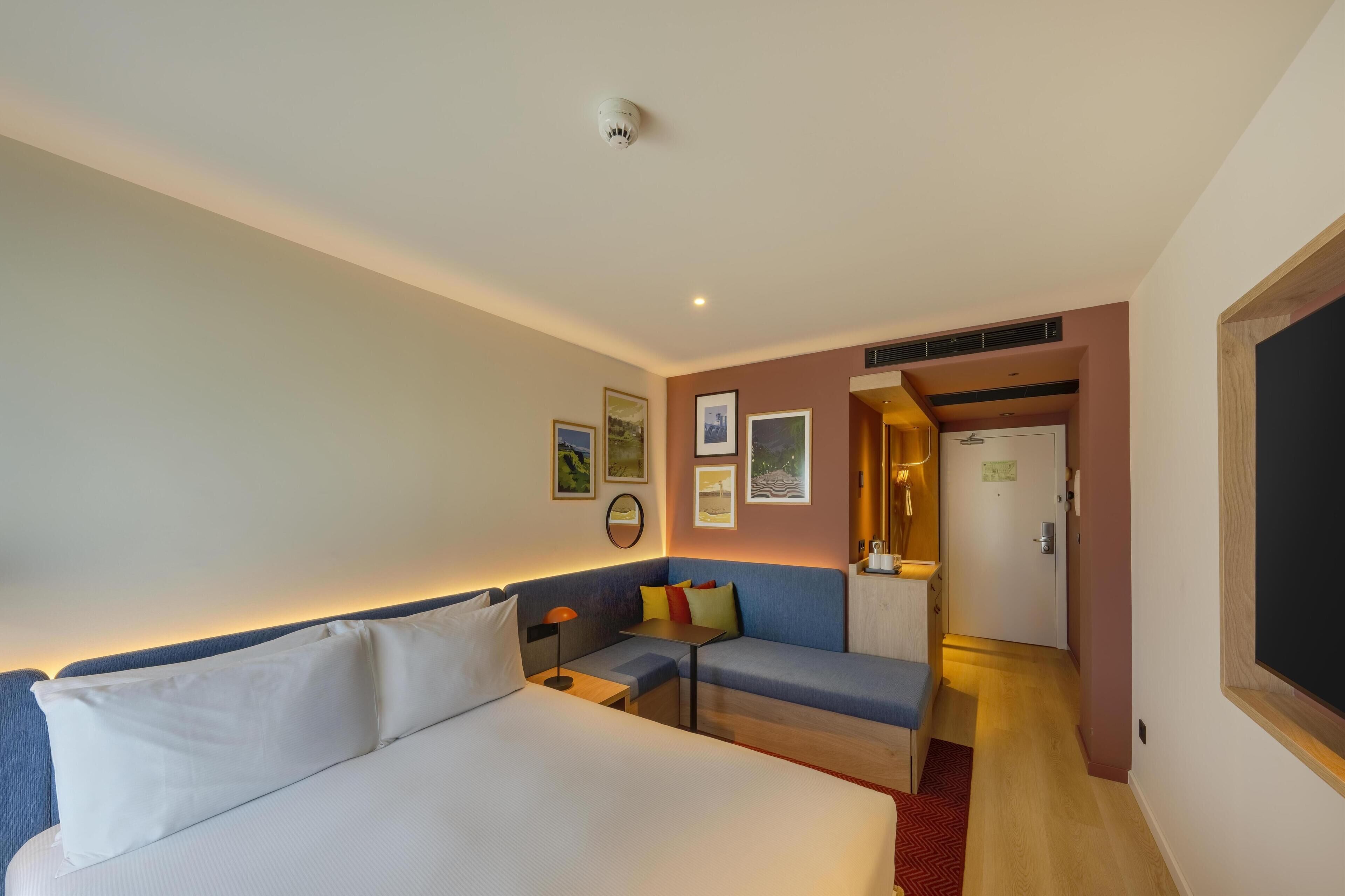 Una camera d'albergo con un letto, un divano, un tavolino e una porta che dà su un corridoio.