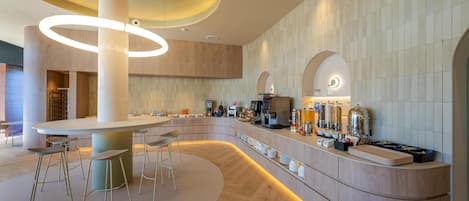 A modern, spacious lobby with a central circular table, bar stools, and a long counter with various amenities.