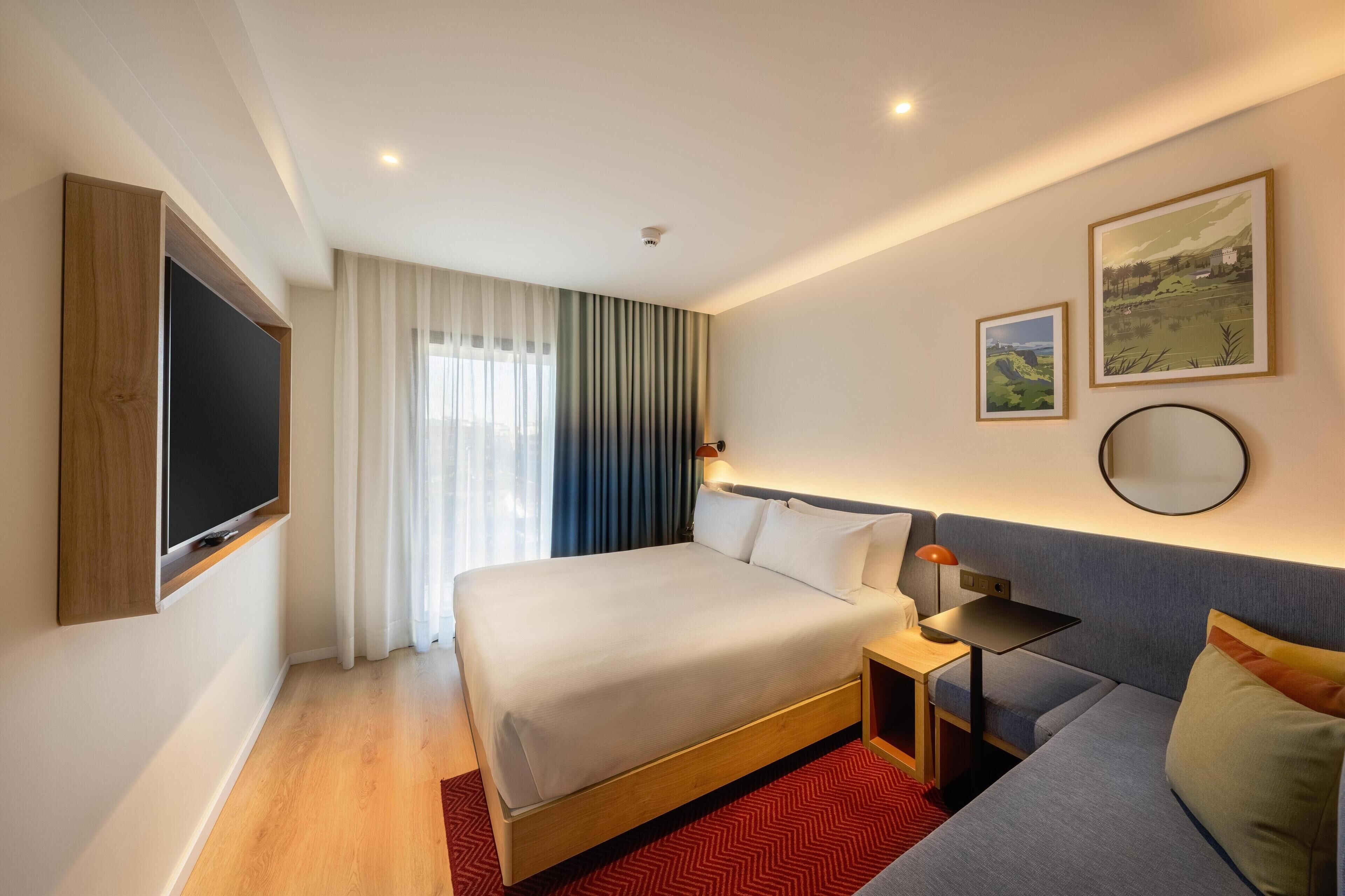 Ein Hotelzimmer mit einem Bett, einem Fernseher, einem Nachttisch mit Lampe, einer Couch und zwei gerahmten Bildern an der Wand.