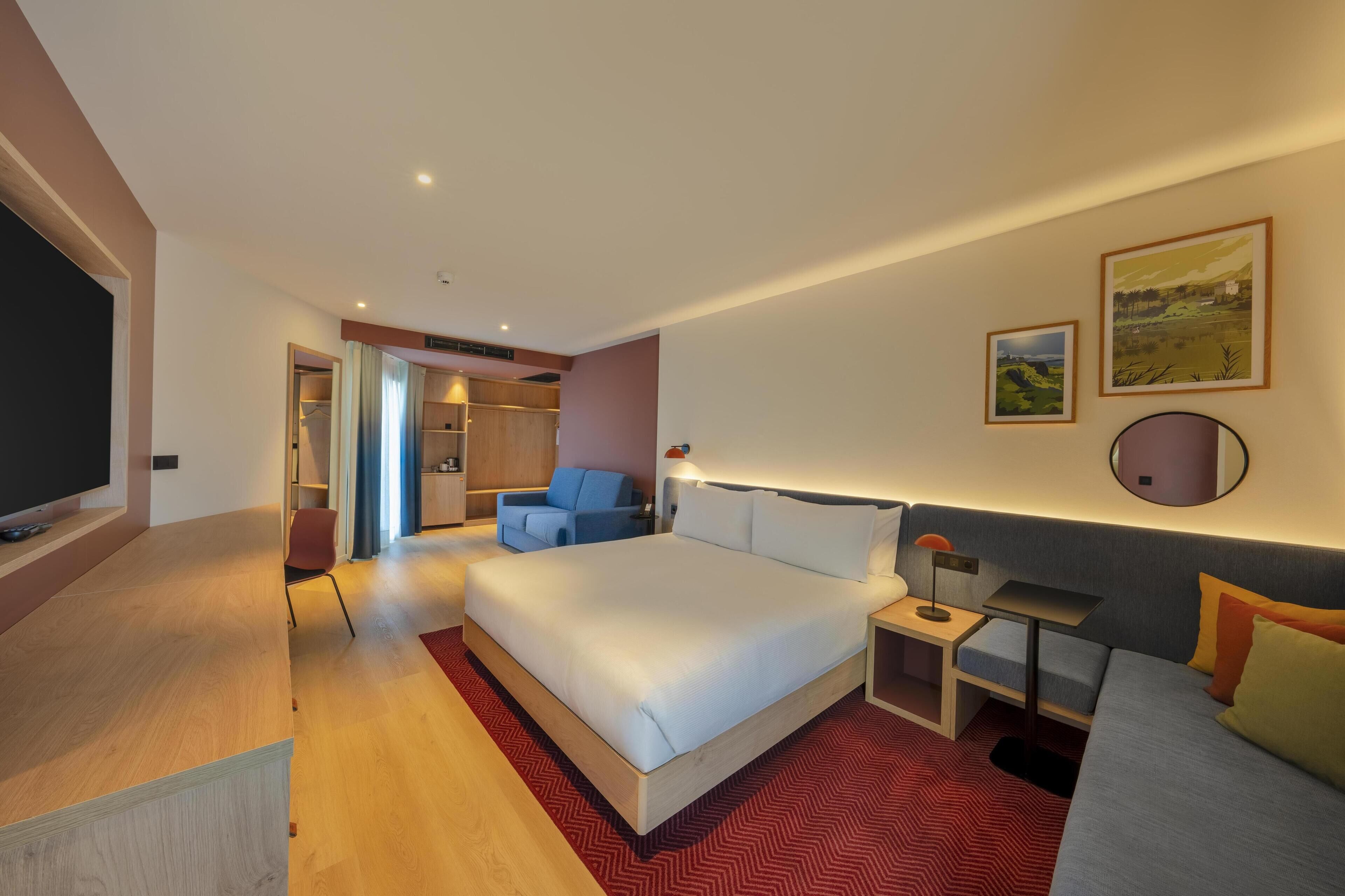Moderni hotellihuone, jossa on suuri sänky, televisio, työpöytä, tuoli ja sohva.