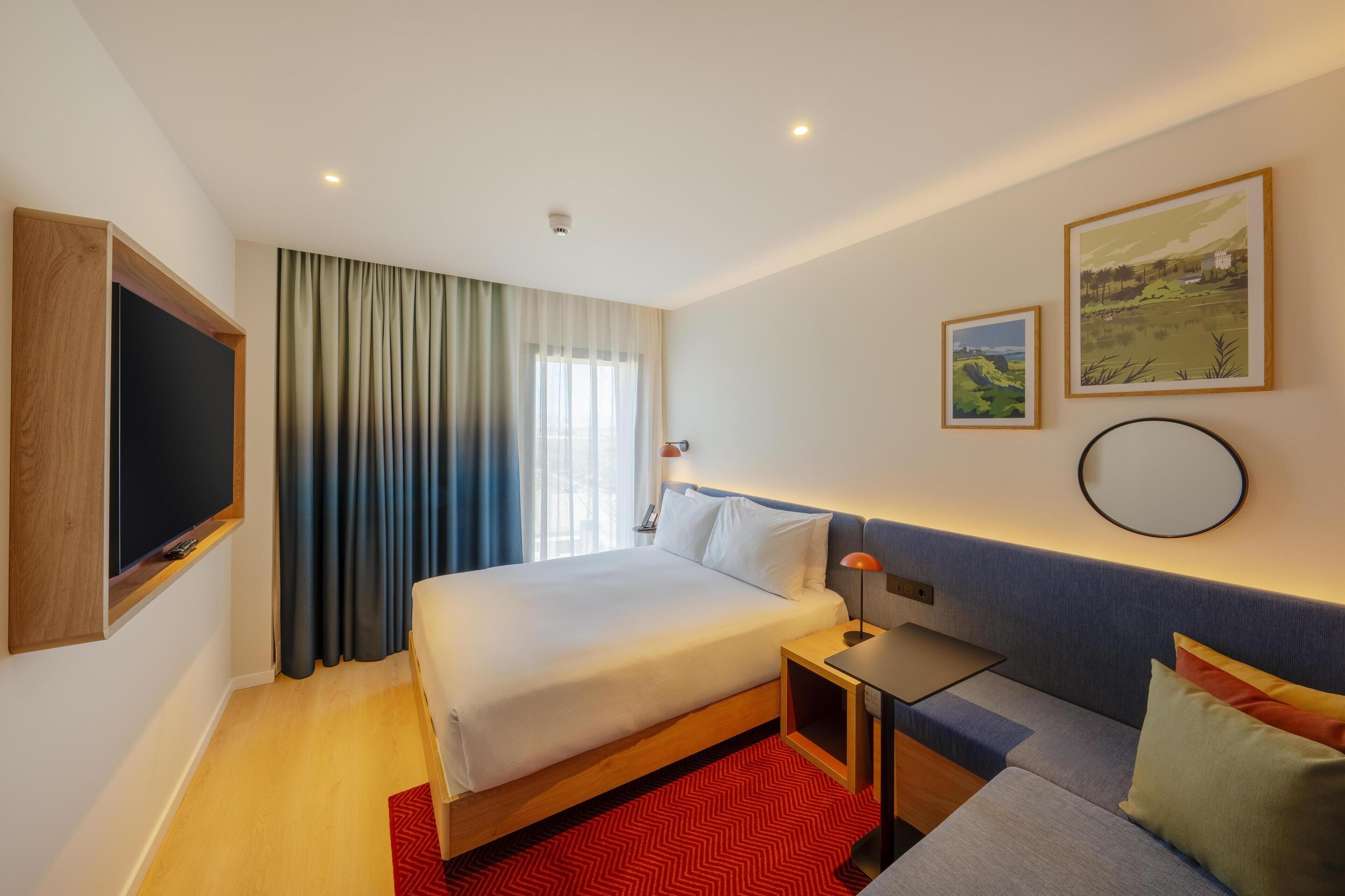 Una habitación de hotel con cama, mesita de noche, lámpara y un ventanal con cortinas.