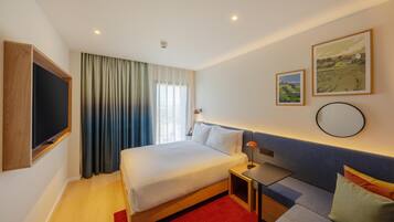 Una habitación de hotel con cama, mesita de noche, lámpara y un ventanal con cortinas.