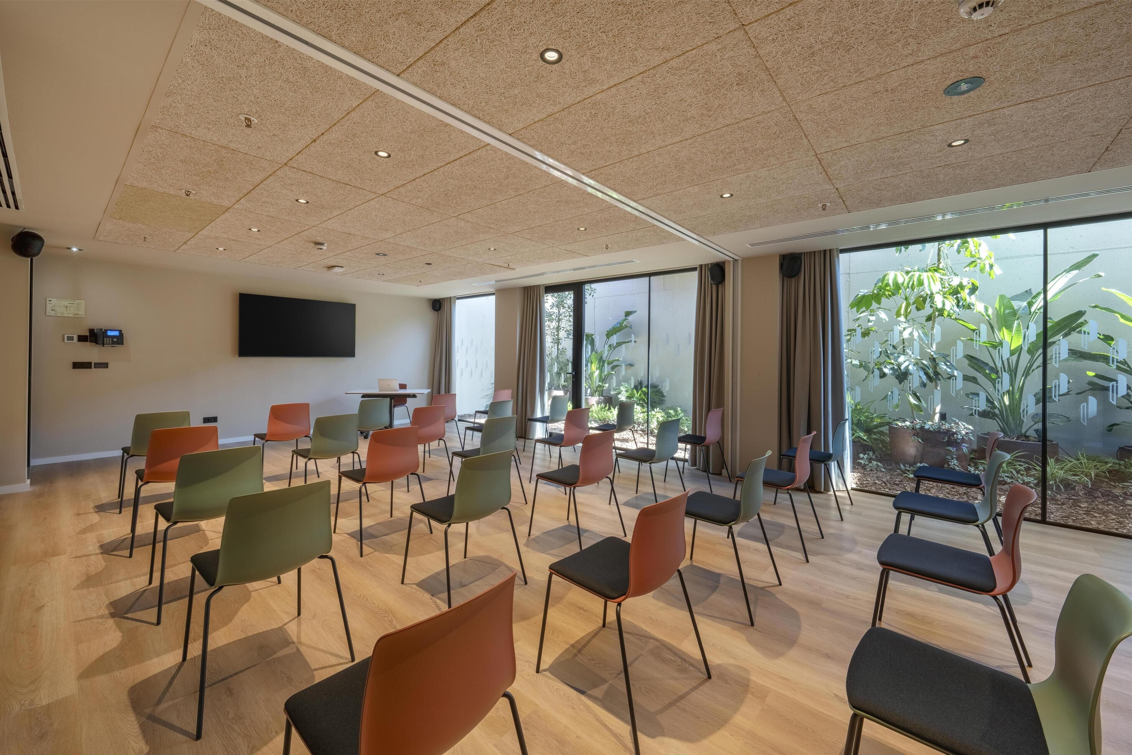 Une salle de conférence équipée d’un téléviseur à écran plat, de plusieurs chaises et de grandes fenêtres donnant sur des plantes.
