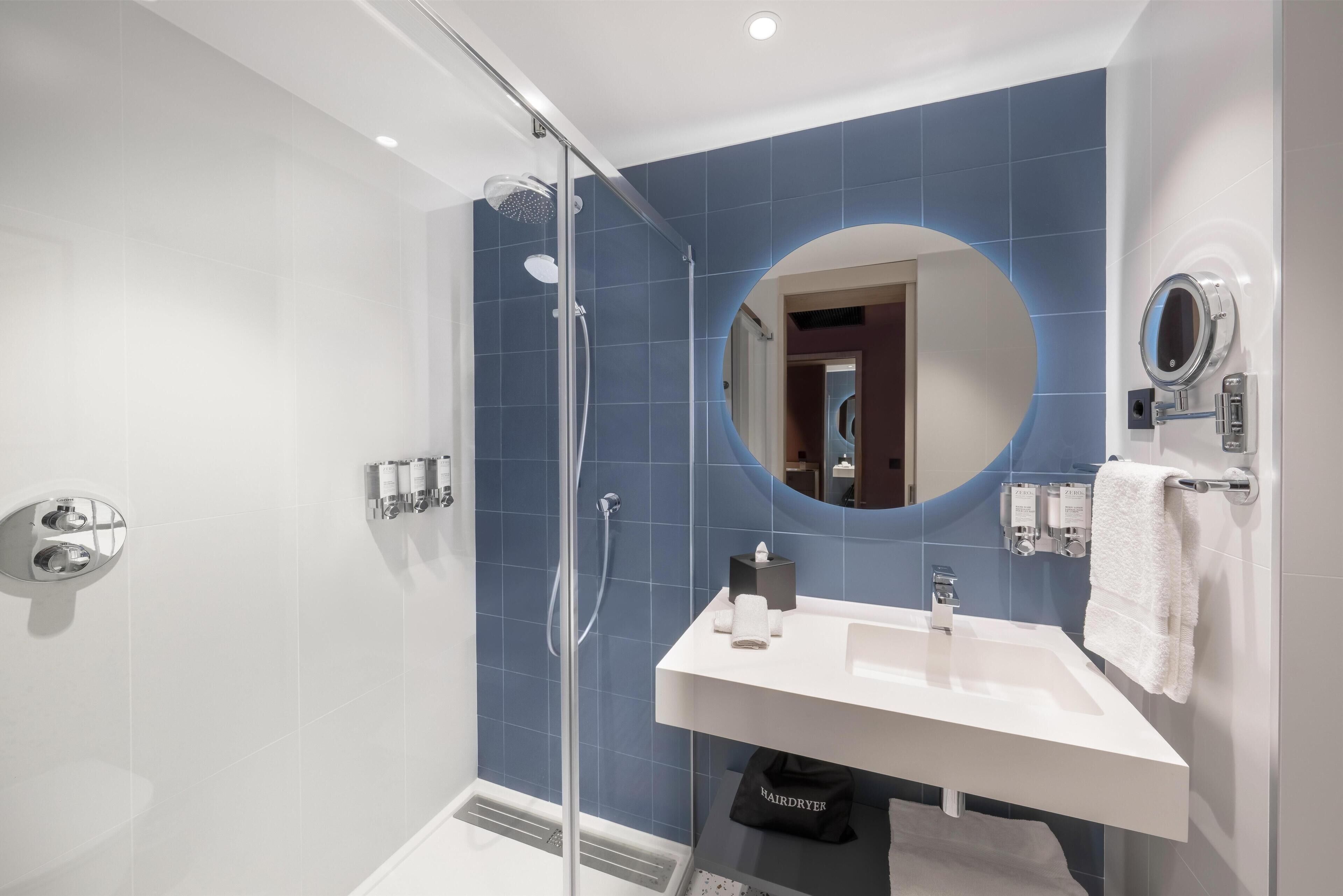 A modern bathroom with a glass-enclosed shower, a white sink, a round mirror, and a towel rack.