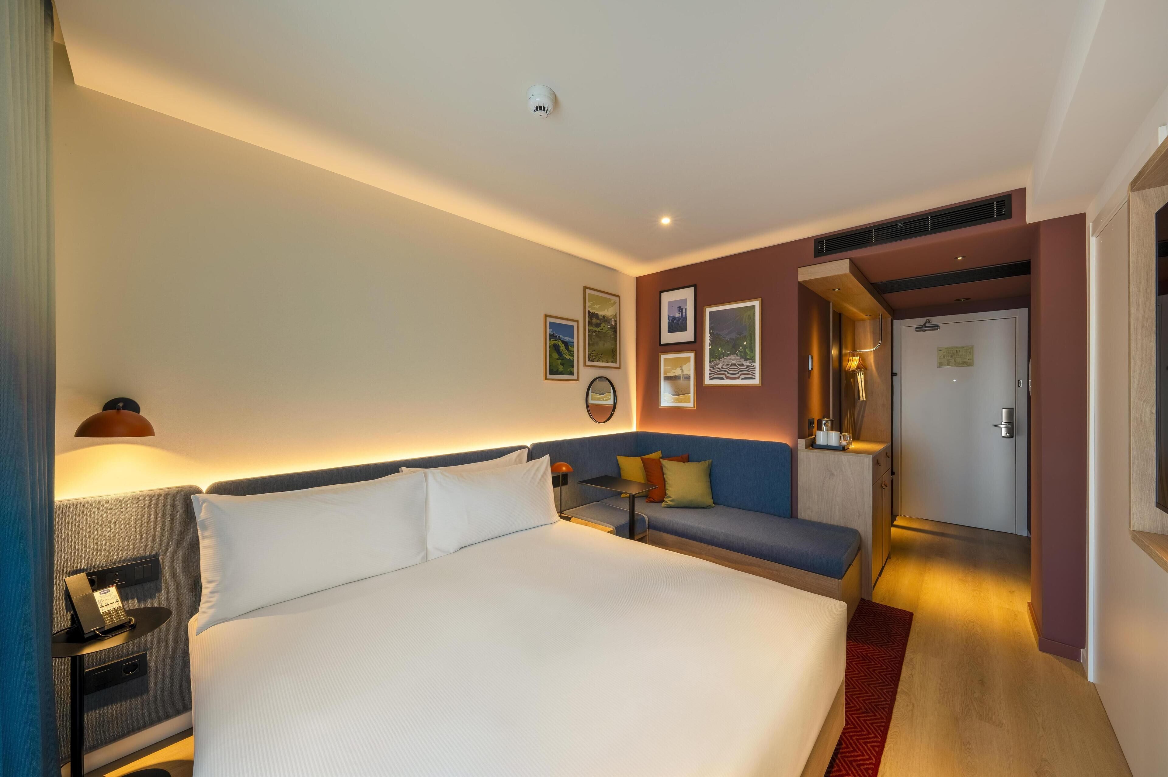 A hotel room with a large bed, a desk with a telephone, a sofa, and a small table with a lamp.