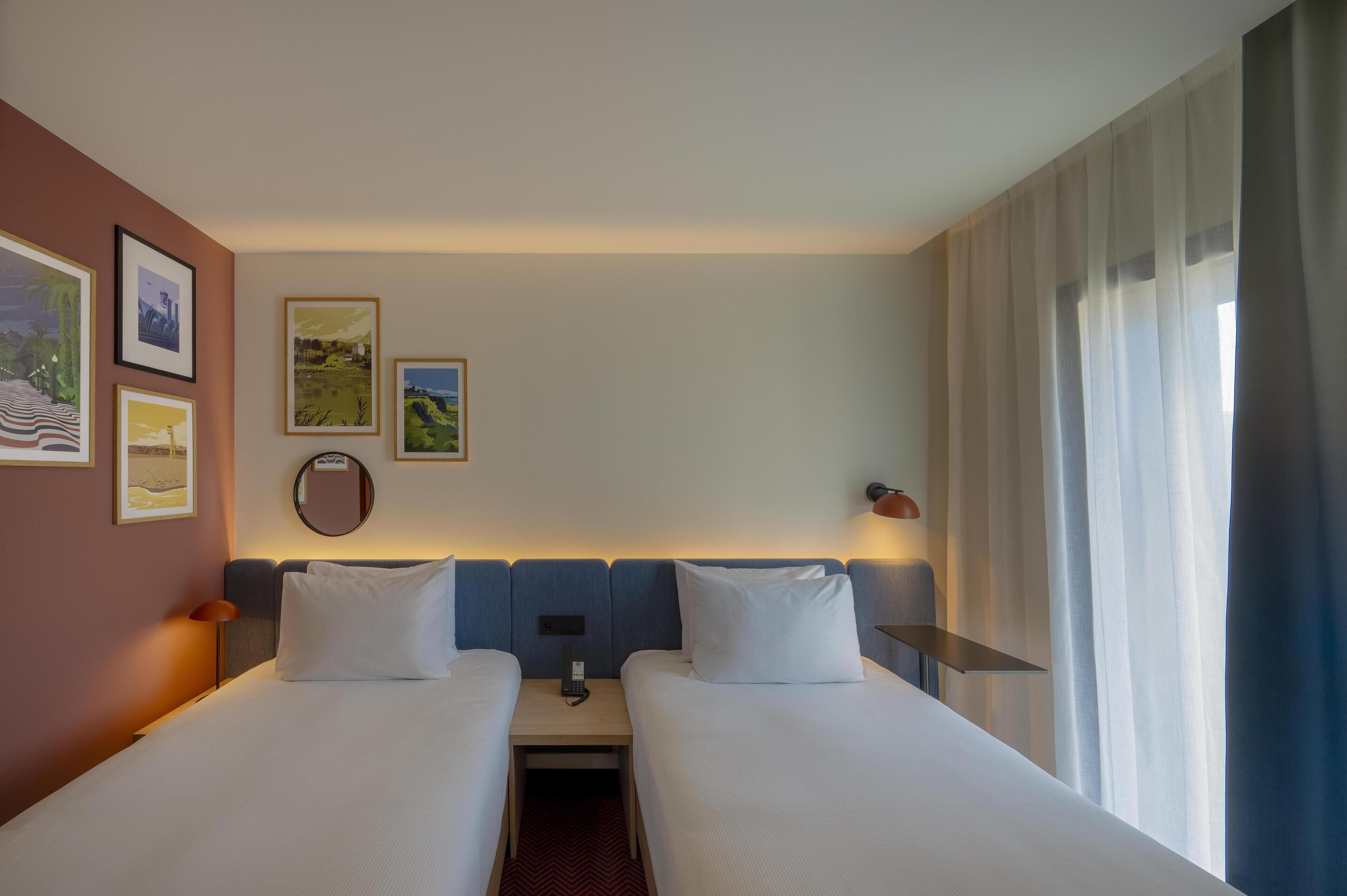 Habitación de hotel con dos camas, una mesita de noche con lámpara, espejo y cuadros en la pared.