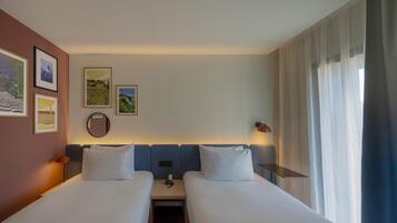 Een hotelkamer met twee bedden, een nachtkastje met een lamp, een spiegel en beelden in lijsten aan de muur.