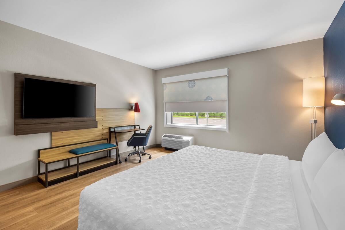 Ein Hotelzimmer mit Bett, Schreibtisch, Stuhl, Fernseher, einem Fenster mit Jalousien und einer Wandlampe.