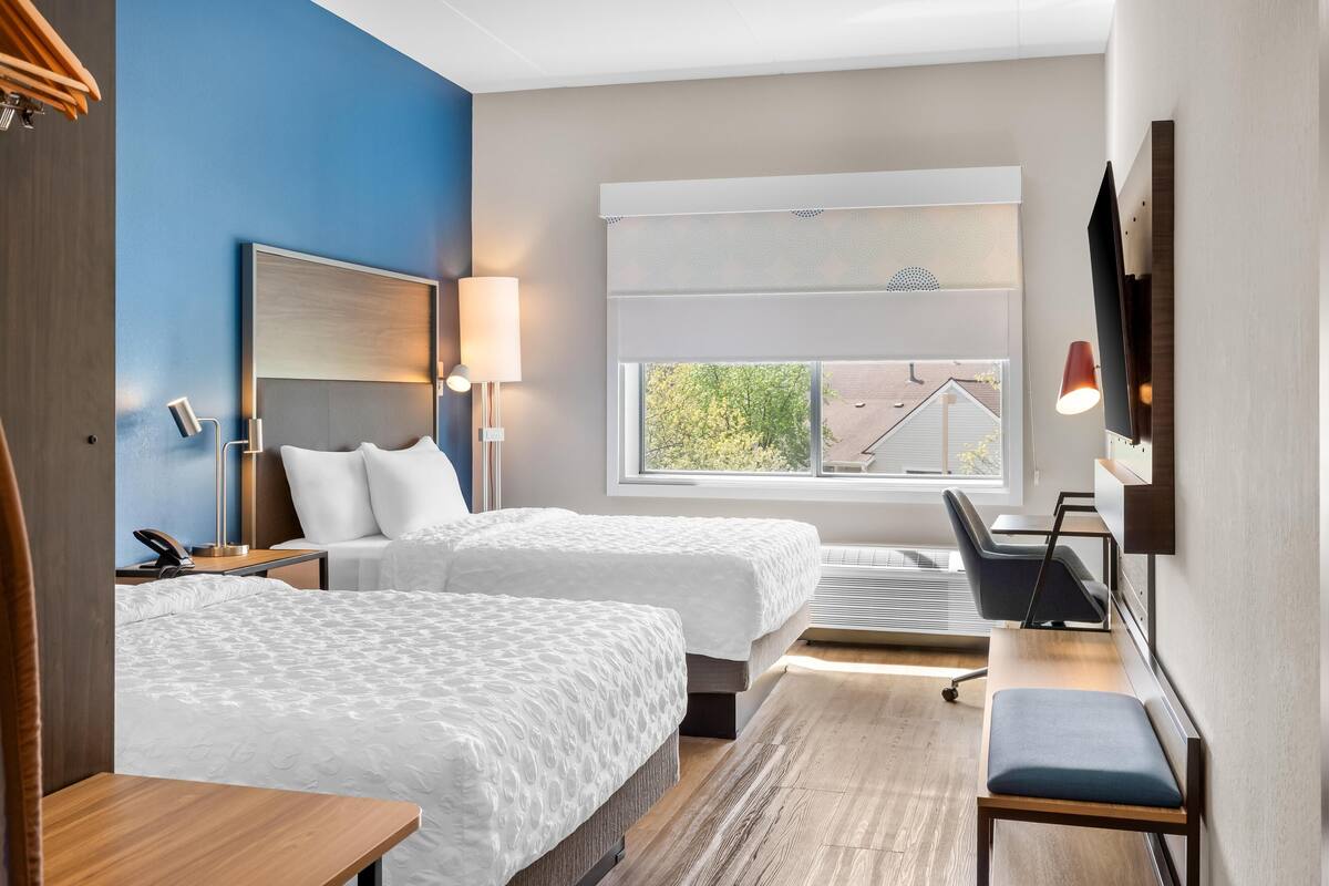 Ein Hotelzimmer mit zwei Betten, einem Schreibtisch, einem Sessel, einem Fernseher und einem Fenster mit Blick auf Bäume.