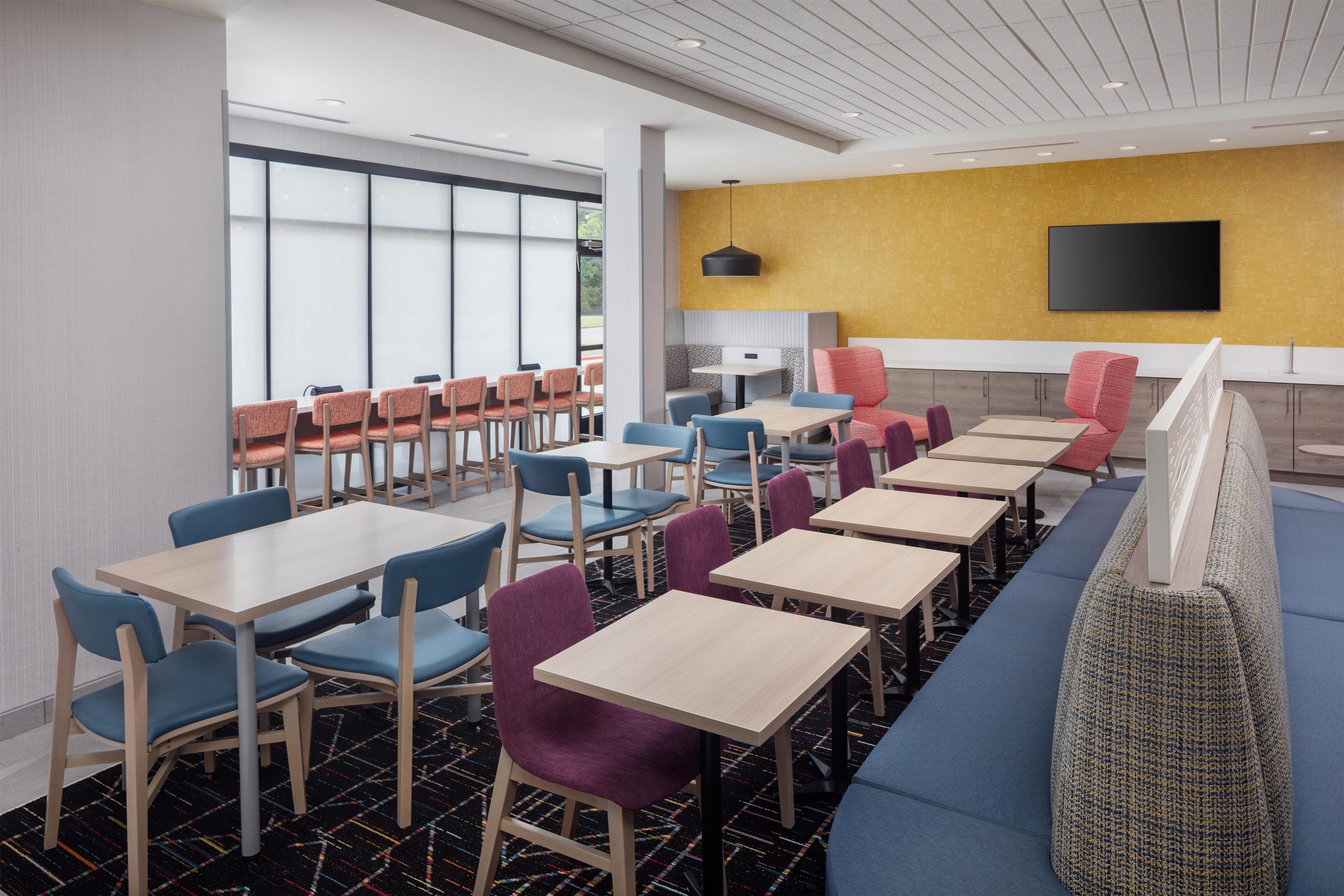 A modern restaurant with multiple tables and chairs, a large window, and a flat-screen TV mounted on the wall.